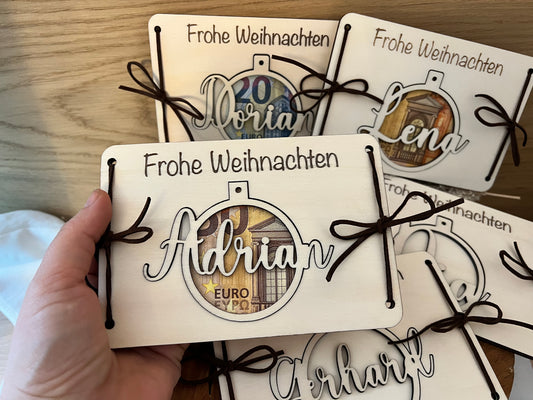 Personalisiertes Geldgeschenk in einer Weihnachtskugel. Das Geld wird zwischen den 2 Holzkarten eingelegt. Die Weihnachtskugel wird mit deinem Wunschnamen personalisiert. Damit kann man auch den Weihnachtsbaum schmücken.