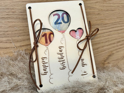 Geldgeschenk Happy Birthday. Gravierte Holzkarte mit der Aufschrift Happy Birthday to you und ausgeschnittenen Luftballons, wo das Geld durchseint.